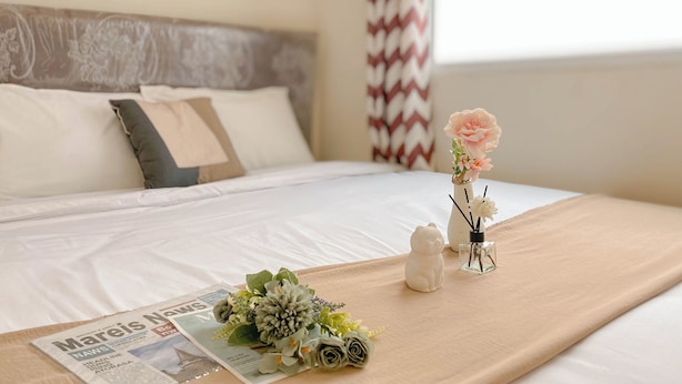 A neatly made bed with a beige bedspread, a floral arrangement, a small cat figurine, and a newspaper on the bed.