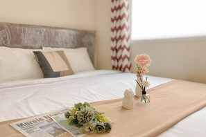 A neatly made bed with a beige bedspread, a floral arrangement, a small cat figurine, and a newspaper on the bed. - Comfort Living 2Br Green Pramuka City Near Mall Apartment (Jakarta)