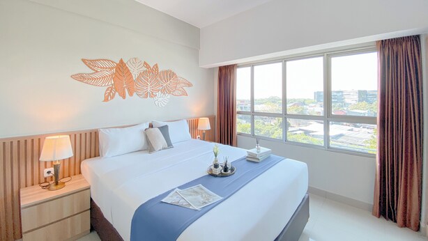 A hotel room with a large bed, a nightstand with a lamp, a decorative wall piece, and a window with curtains.