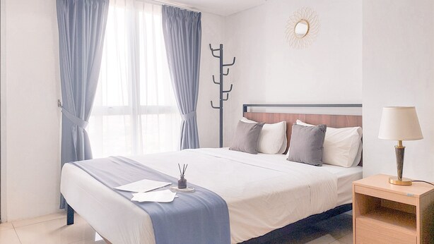 Una cama bien hecha con una colcha azul, una mesita de noche con una lámpara y un perchero apoyado en la pared.