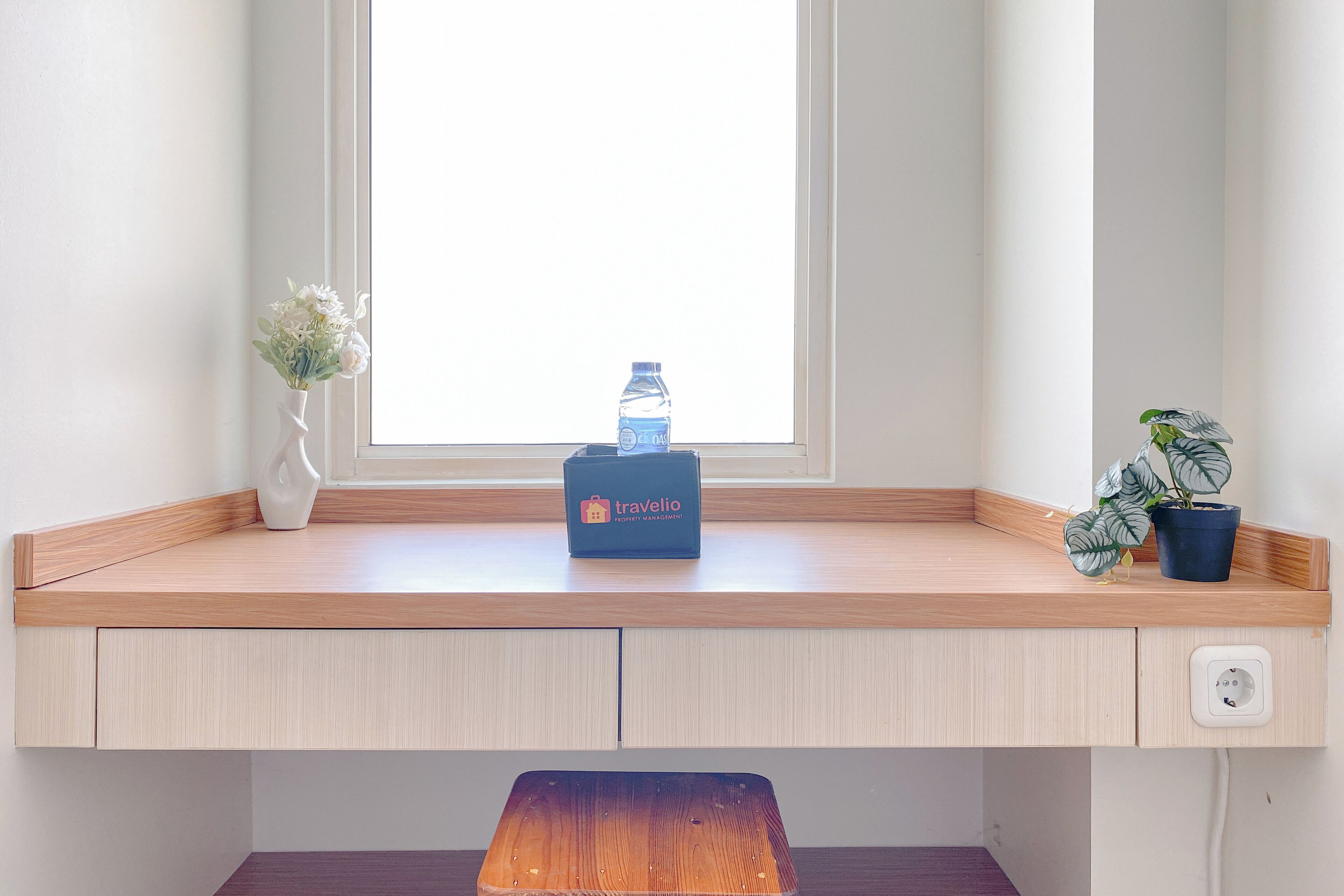 A corner desk with a wooden surface, a small vase with flowers, a water bottle, a potted plant, and a travel bag.