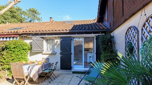Une terrasse avec une table et des chaises, un petit jardin et un bâtiment doté d’un toit en tuiles et de volets en bois.