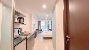 A compact hotel room with a kitchenette, a bed, a desk, and a window with a view of trees. - Modern Look Studio (No Kitchen) Apartment At Skales Residence Nusa Dua Bali (Nusa Dua)
