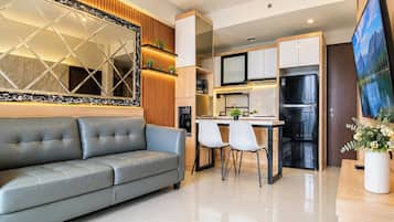 A modern living room with a grey sofa, a dining area with two chairs, a kitchen with a refrigerator, and a large mirror.