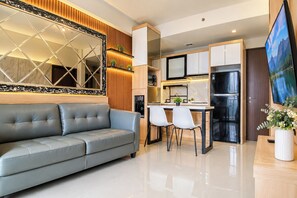 A modern living room with a grey sofa, a dining area with two chairs, a kitchen with a refrigerator, and a large mirror. - Cozy And Good Place 2Br Transpark Cibubur Apartment (Depok)