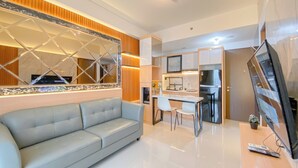 A modern living room with a grey sofa, a dining area with a white table and chairs, a kitchen with a TV, and a large mirror. - Cozy And Good Place 2Br Transpark Cibubur Apartment (Depok)