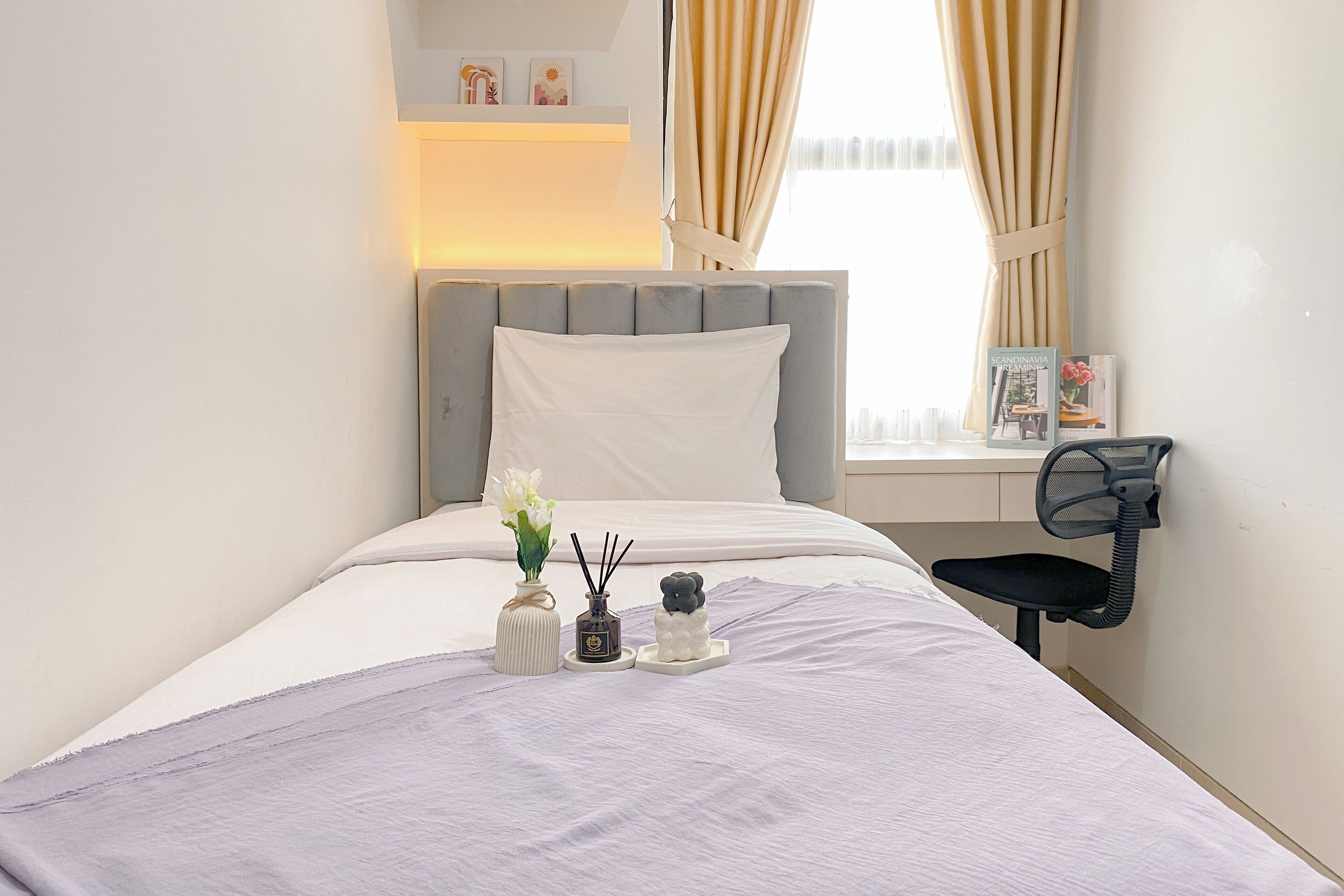 A neatly made bed with a grey headboard, a bedside table with a lamp, and a window with curtains.