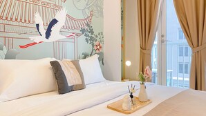 A hotel room with a bed, a decorative wall mural, a vase with flowers, and a view of the city through the window. - Affordable And Homey Studio At Tokyo Riverside Pik 2 Apartment (Teluknaga)