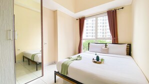 A hotel room with a bed, a window with curtains, a view of buildings outside, and a small table with a vase and a plant. - Minimalist And Cozy 1Br Casa De Parco Apartment (Pagedangan)