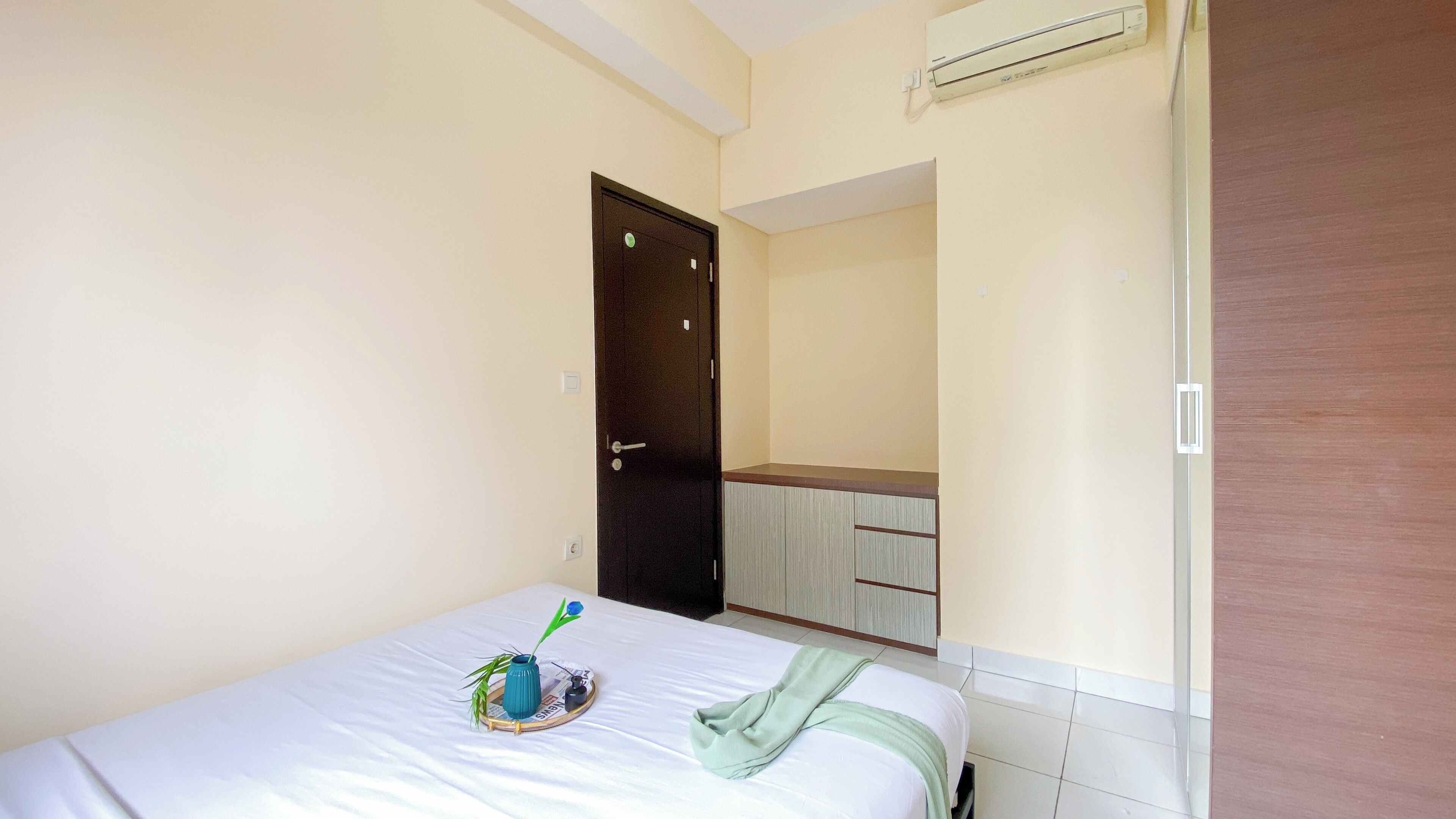 A hotel room with a bed, a small table with a vase and a newspaper, a mirror, and a door.