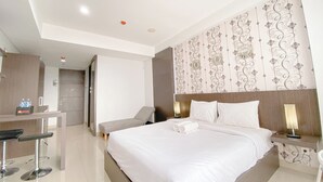 A hotel room with a large bed, a desk with a chair, a bench, and a bathroom with a mirror. - Tranquil Studio Room At Dago Suites Apartment (Bandung)
