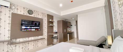 A hotel room with a large bed, a TV mounted on the wall, a small kitchenette, and a bathroom visible through an open door.