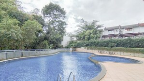 A swimming pool with a curved design, surrounded by a fence and trees. There are lounge chairs and a building in the background. - Tranquil Studio Room At Dago Suites Apartment (Bandung)