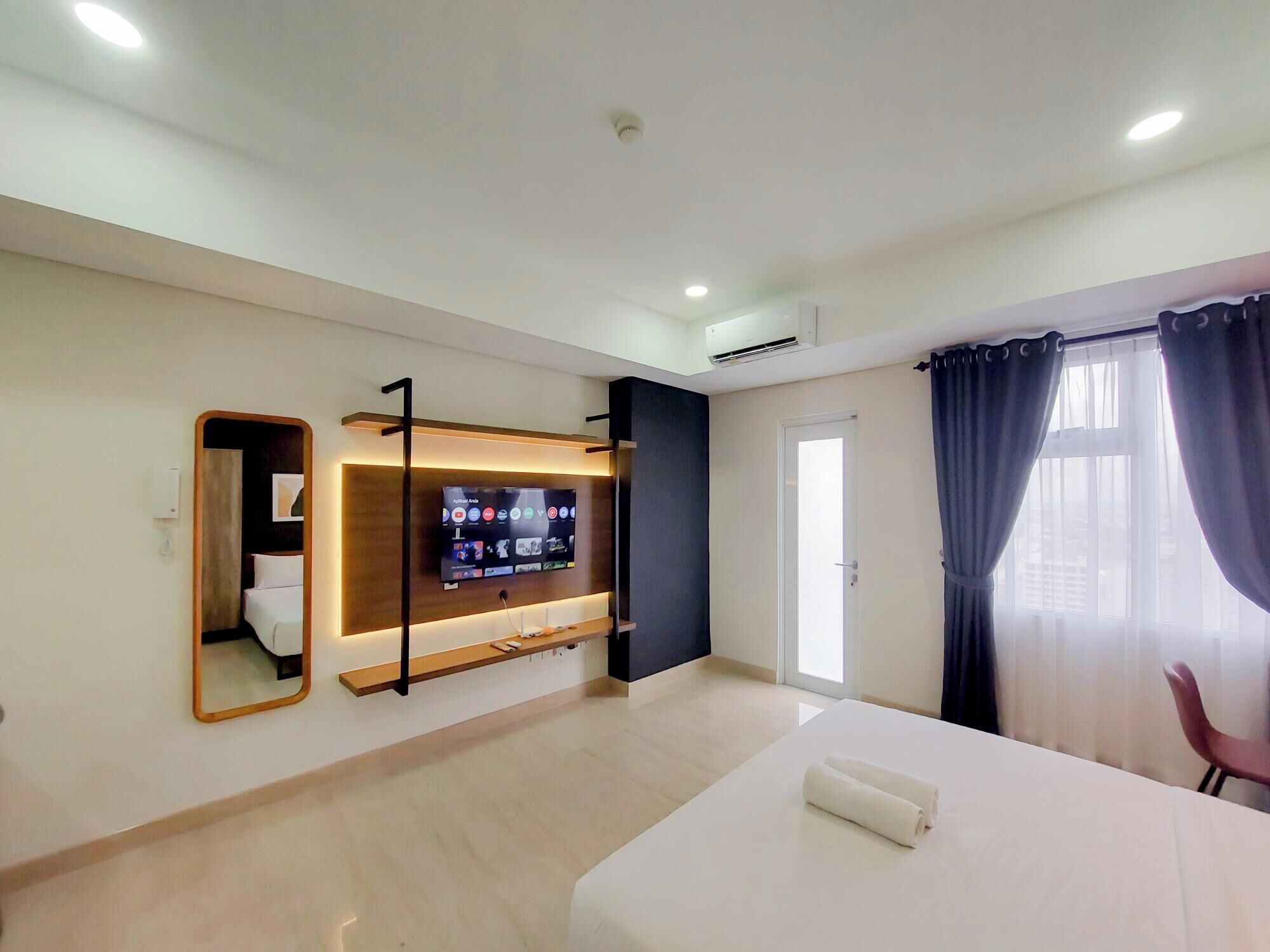 A modern hotel room with a flat-screen TV mounted on the wall, a wooden shelf below, a bed with a white mattress, a chair, and a large window with curtains.
