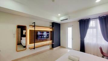 A modern hotel room with a flat-screen TV mounted on the wall, a wooden shelf below, a bed with a white mattress, a chair, and a large window with curtains.