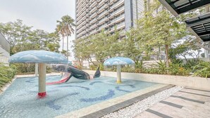 A swimming pool with two slides, surrounded by greenery and a tall building in the background. - Restful And Simple 2Br At The Via And The Vue Apartment (Surabaya)
