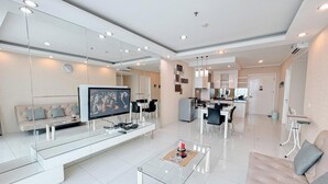 A modern living room with a flat-screen TV, a glass coffee table, a sofa, and a dining area with chairs. - Restful And Simple 2Br At The Via And The Vue Apartment (Surabaya)