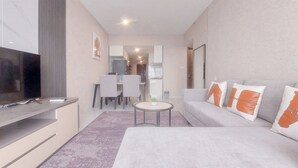 Un soggiorno moderno con una TV a schermo piatto, un divano grigio con cuscini arancioni, un tavolino rotondo con una pianta e un'area pranzo sullo sfondo.