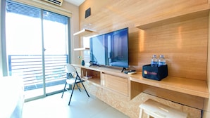 Una camera d'hotel moderna con una grande TV a schermo piatto, una scrivania in legno, una sedia, un tavolino e una panca. Sulla scrivania ci sono due bottiglie d'acqua e sulla panca una borsa da viaggio.