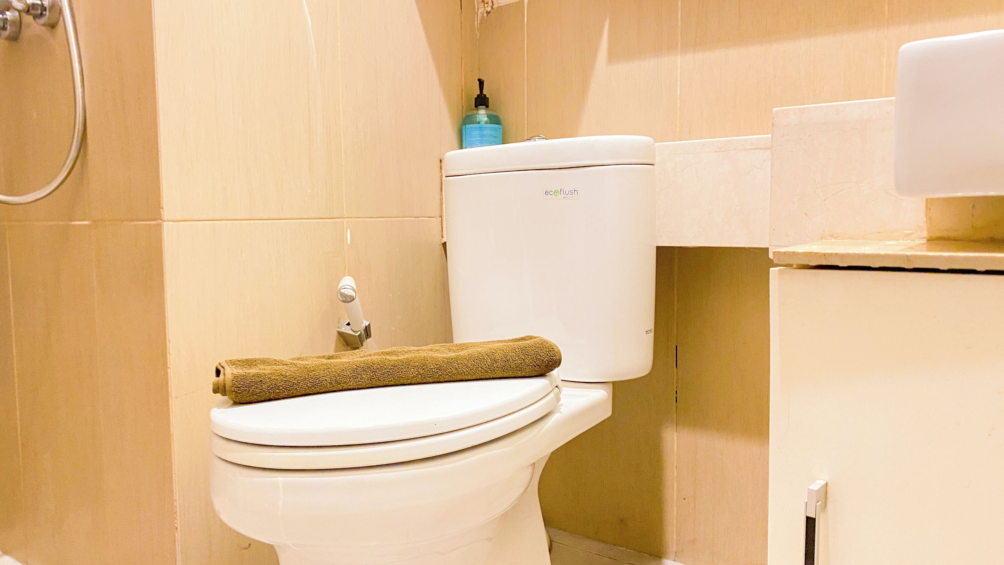 Des toilettes avec une serviette posée dessus, un distributeur de savon et une pomme de douche en arrière-plan.