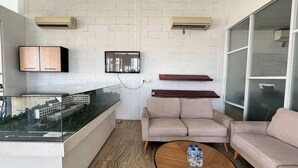 A modern living room with a glass partition, a beige sofa, a coffee table with water bottles, and a mounted TV. - Modern Furnished Studio (No Kitchen) Apartment At Skales Residence Nusa Dua Bali (Nusa Dua)