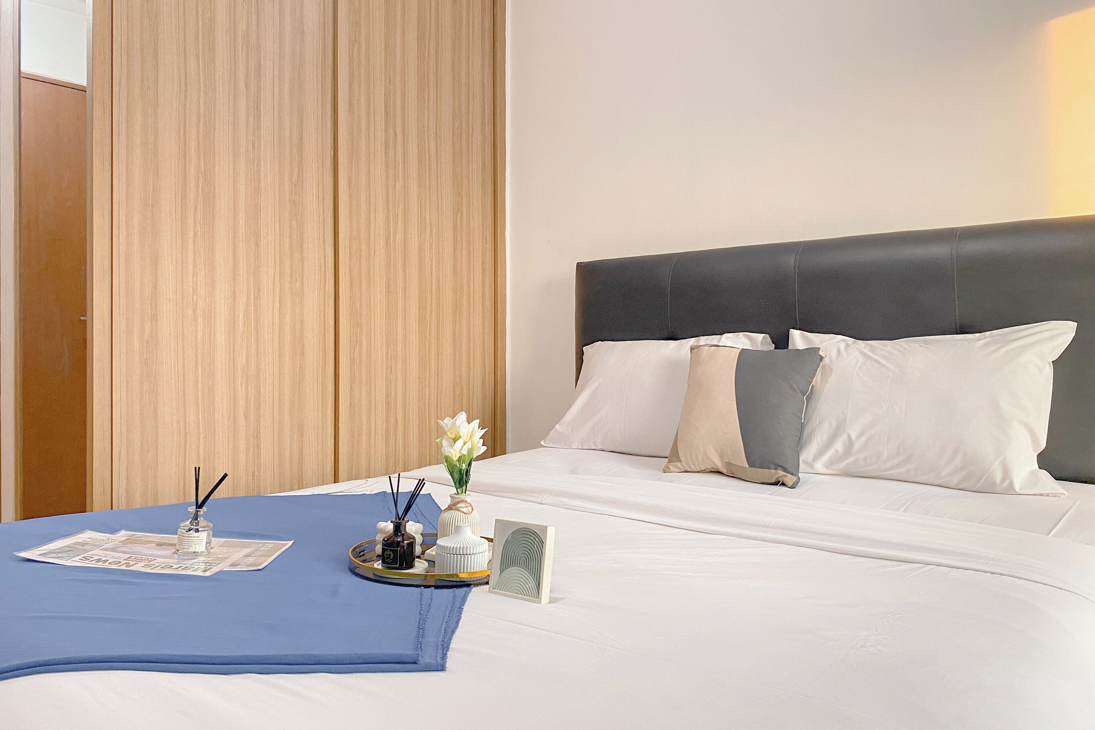 A neatly made bed with white linens, a blue throw blanket, and decorative items including a vase with flowers and a small clock.