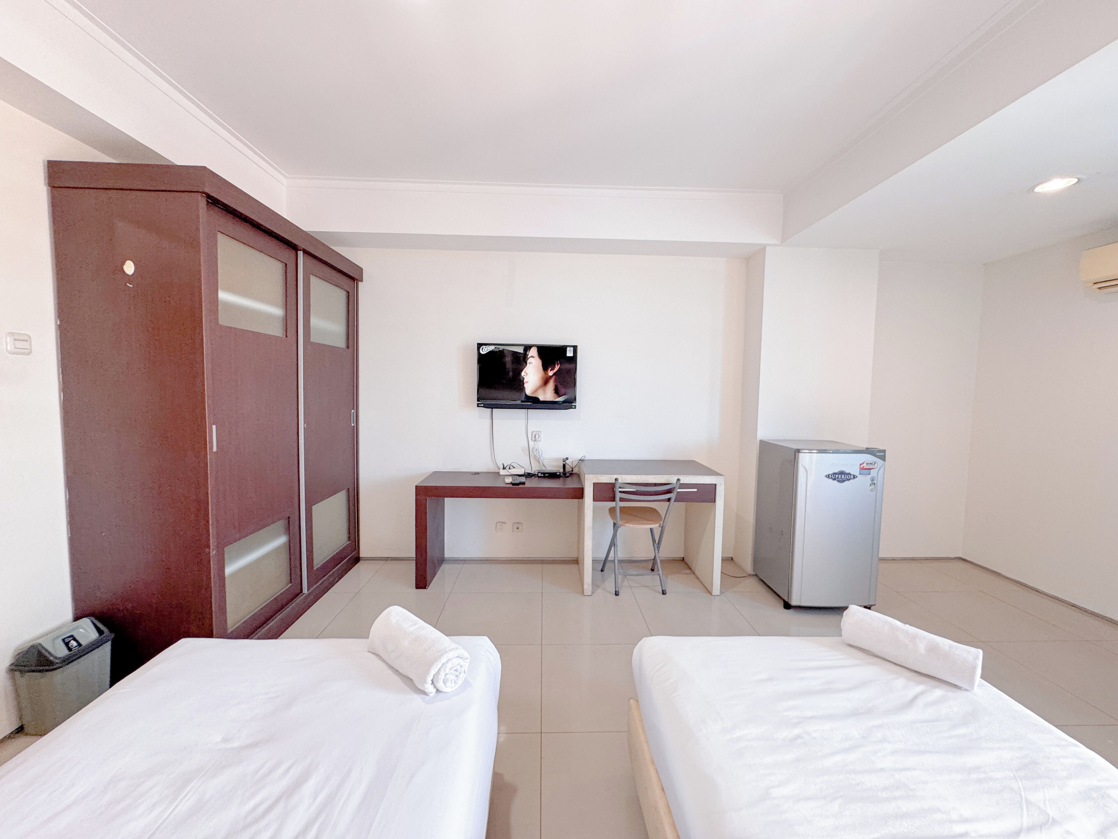 A hotel room with two single beds, a desk with a chair, a television, and a refrigerator.