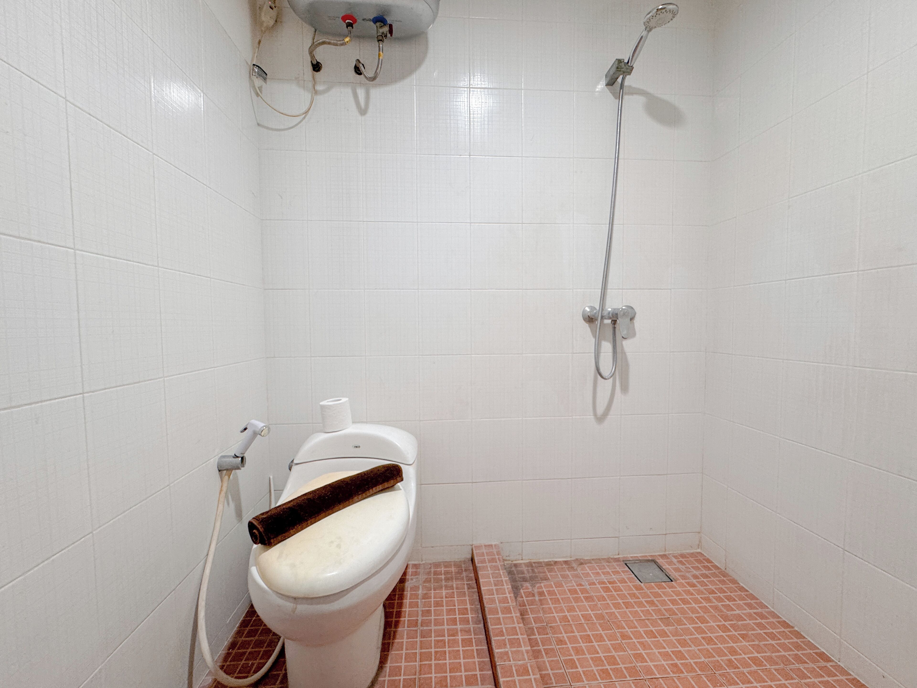 A small, white-tiled bathroom with a toilet, a shower, and a visible pipe.