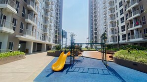 A playground with a yellow slide and exercise equipment in a residential area with high-rise buildings. - Best Price And Comfy Studio At Tokyo Riverside Pik 2 Apartment (Teluknaga)