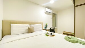 A hotel room with a bed, a desk, a chair, an air conditioner, and a small plant on the bedside table.