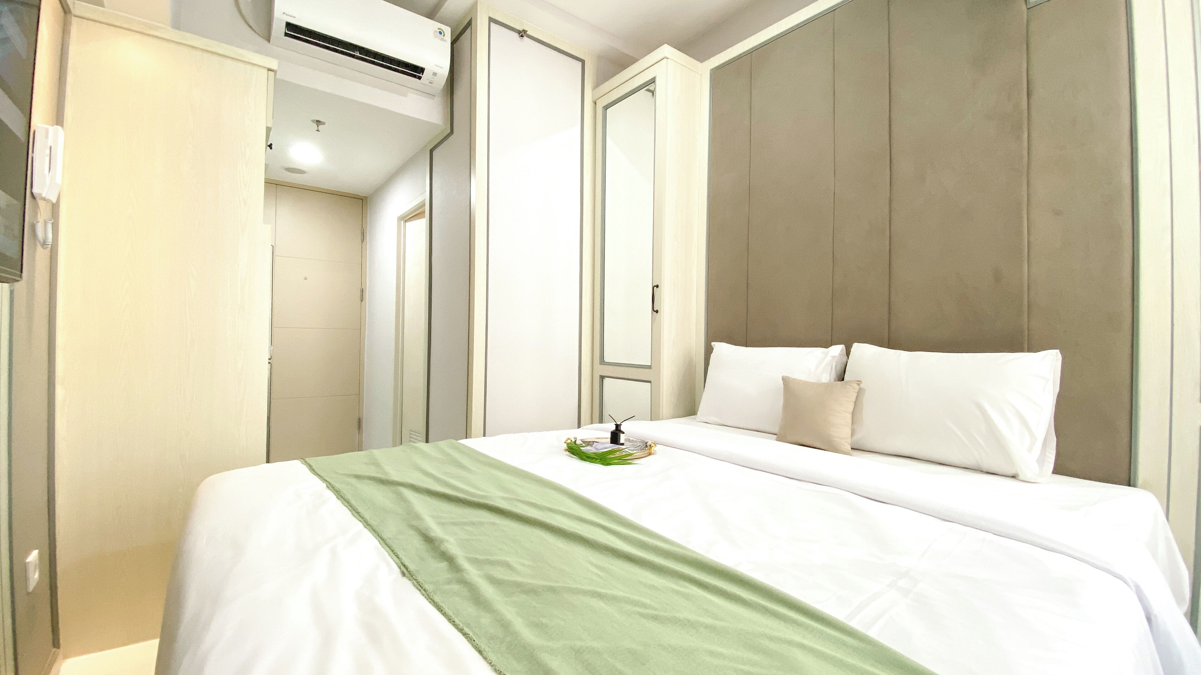 A hotel room with a bed, a green throw, a bedside table, and a wall-mounted air conditioner.