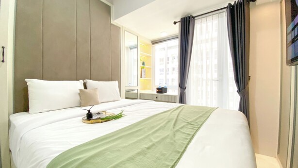 A hotel room with a large bed, a bedside table, a mirror, and a window with curtains. - Cozy And Best Studio Apartment Tokyo Riverside Pik 2 (Teluknaga)