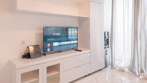 A modern living room with a flat-screen TV on a white cabinet, a small table with a decorative item, and a water dispenser. - Nice Studio Apartment At Sky House Bsd Near Aeon Mall (Pagedangan)