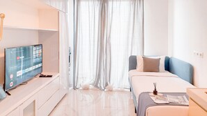 A modern hotel room with a bed, a TV on a stand, and a window with sheer curtains. - Nice Studio Apartment At Sky House Bsd Near Aeon Mall (Pagedangan)