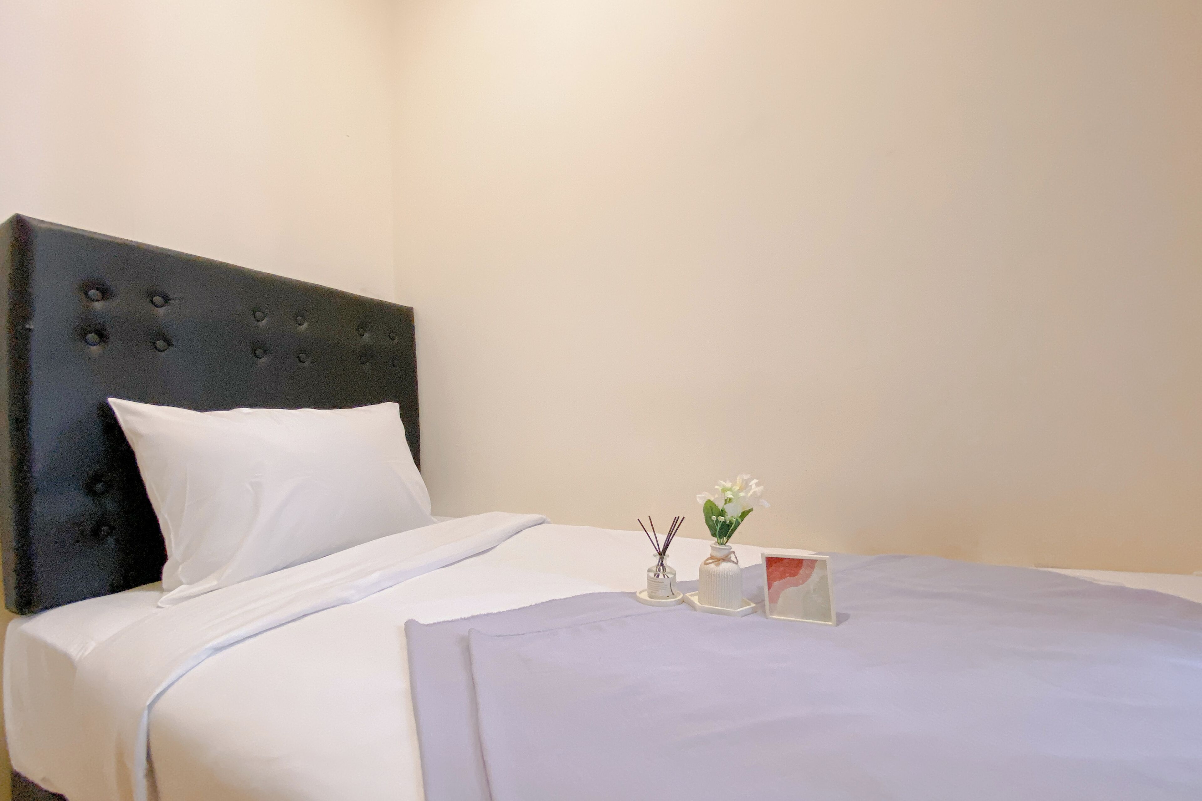 Una cama bien hecha con sábanas blancas, cabecera oscura y una mesita de noche con un jarrón de flores y una vela.