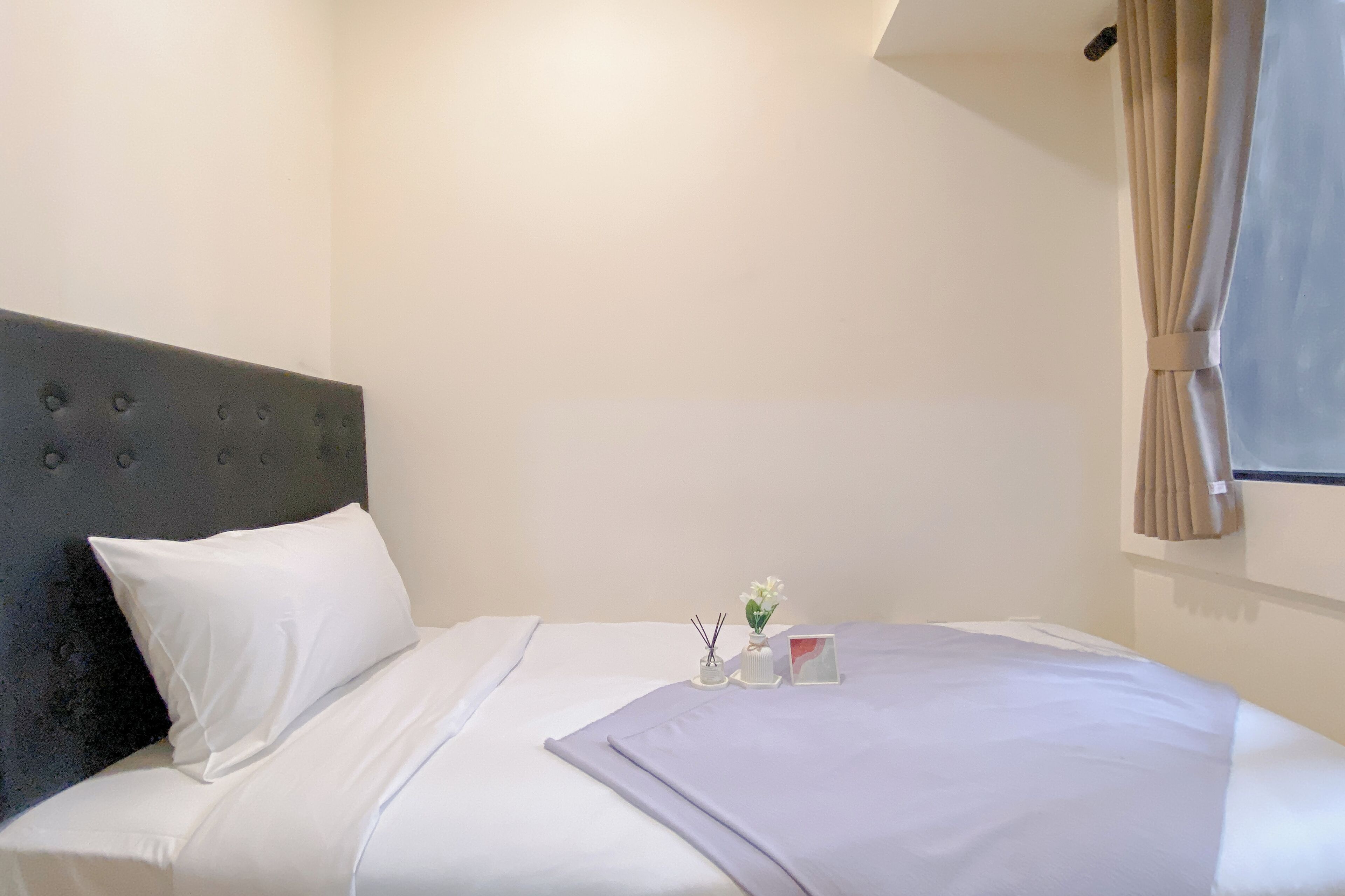 A neatly made bed with white linens, a grey headboard, and a small bedside table with a vase of flowers and a red book.