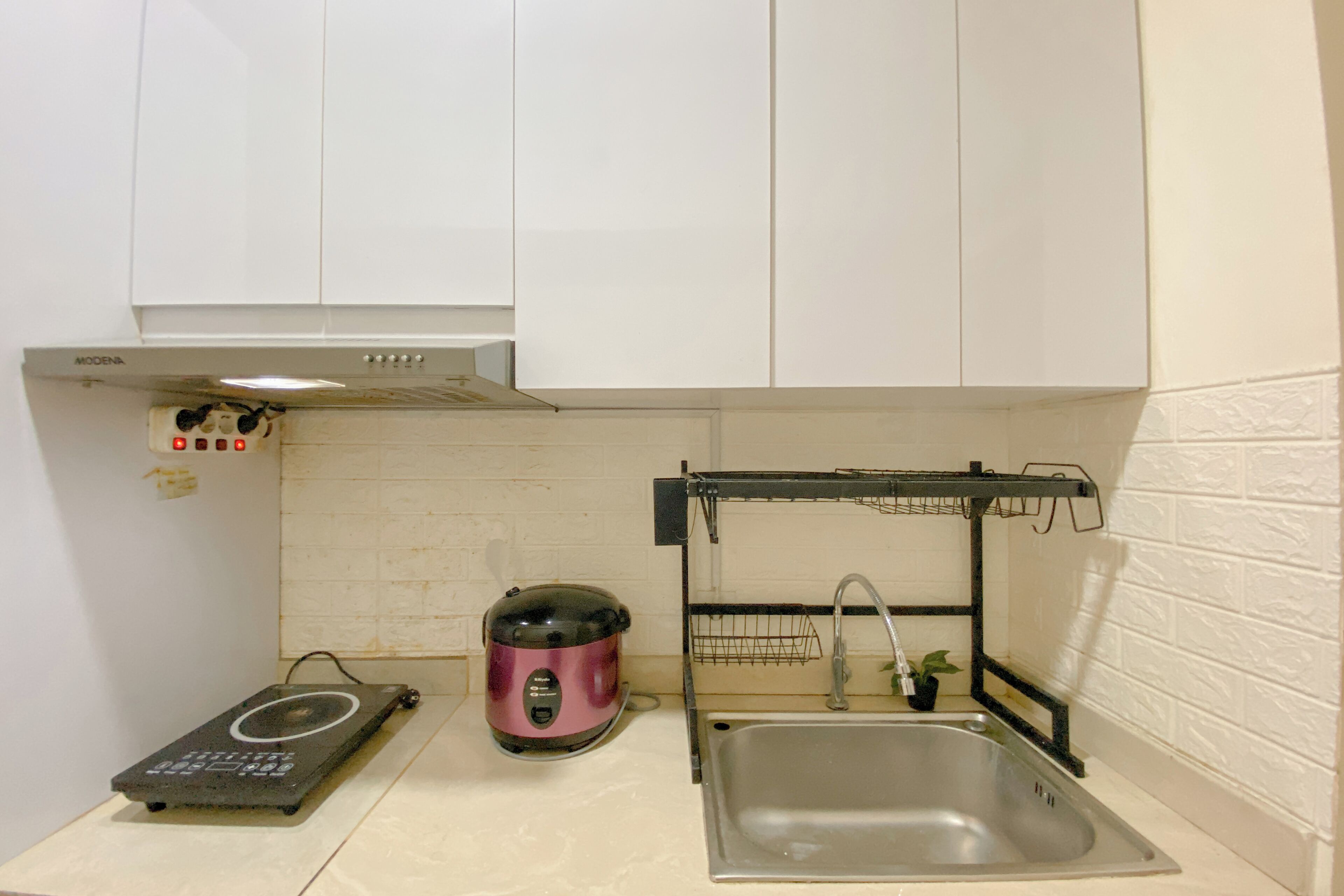 Une cuisine compacte avec des meubles blancs, un évier en acier inoxydable, une plaque de cuisson à induction noire et un cuiseur à riz électrique rose.