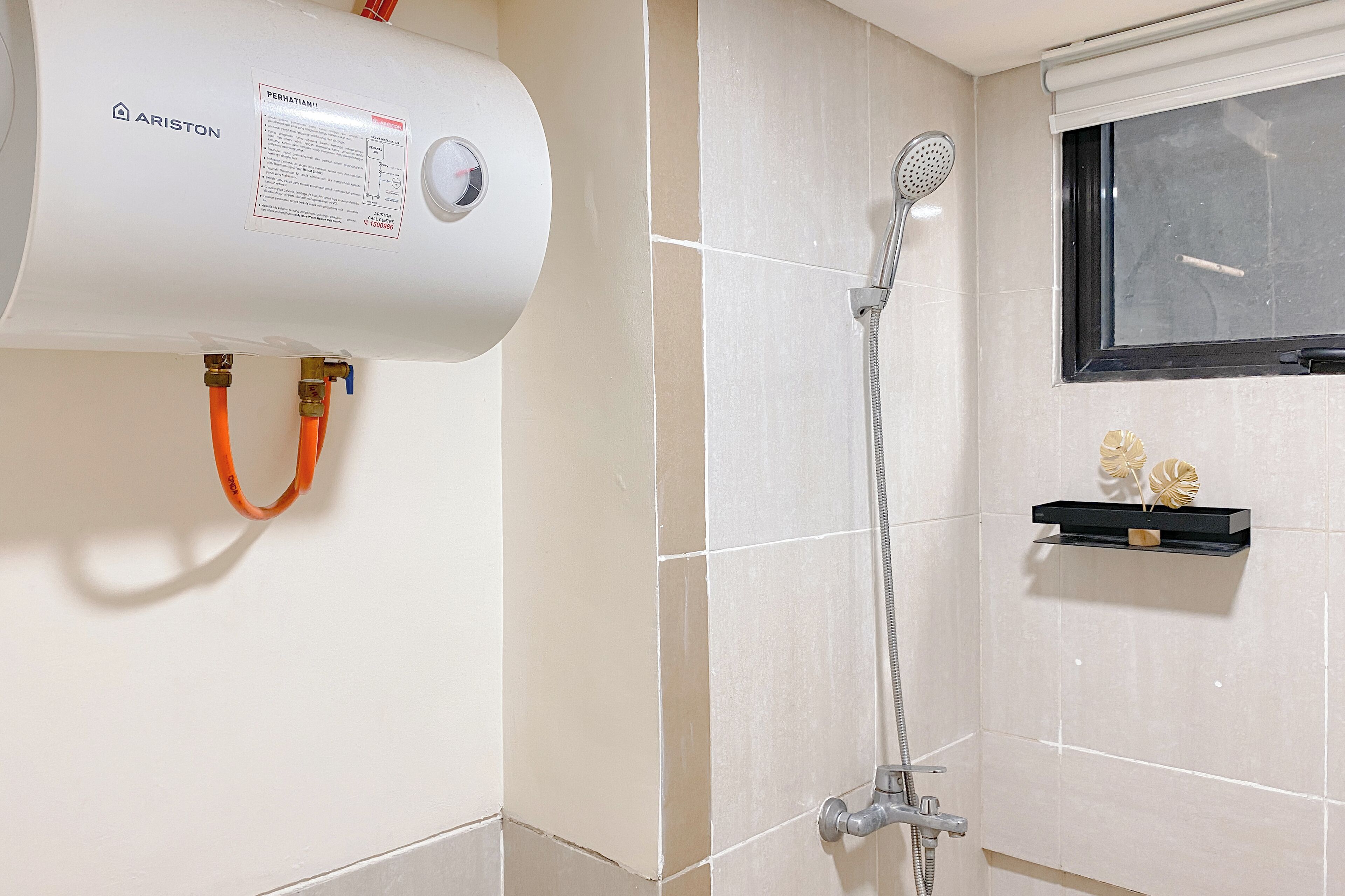 Un baño con pared de azulejos blancos, ducha con cabezal de ducha manual y calentador de agua marca ARISTON.