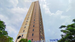 A tall, beige-colored building with multiple windows, surrounded by trees and a partly cloudy sky. - The Stylish Studio At Tamansari Panoramic Apartment (Bandung)