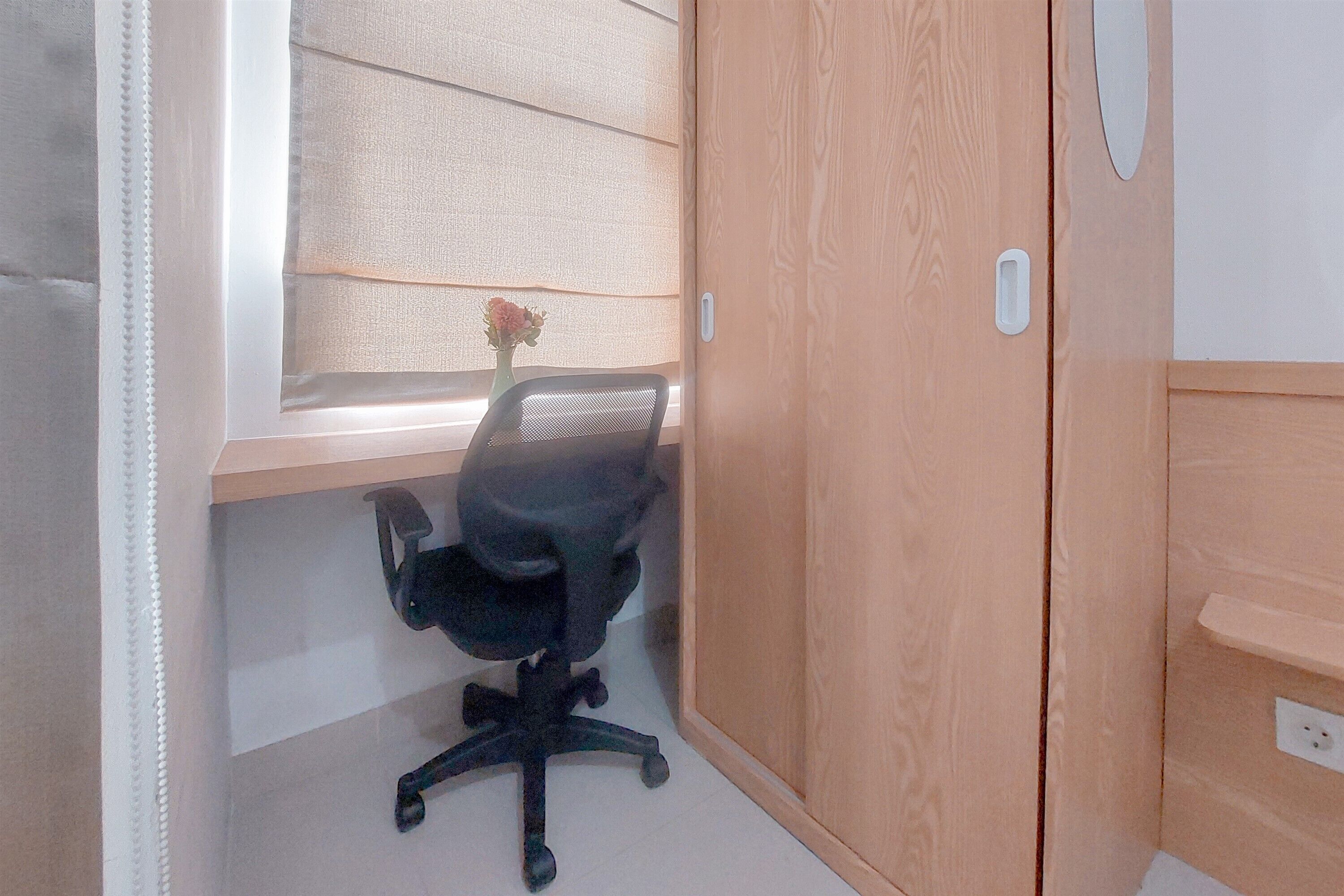 Une pièce avec une armoire en bois, un fauteuil de bureau noir, une petite table et une fenêtre avec des stores.