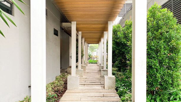 Een overdekte gang met betonnen trappen en witte zuilen, omzoomd door groen en een gebouw aan één kant.