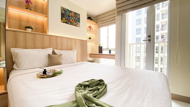 A hotel room with a large bed, a bedside table, a window with a view of buildings, and a wall with a clock and artwork. - Brand New Studio Room At Tokyo Riverside Pik 2 Apartment (Teluknaga)