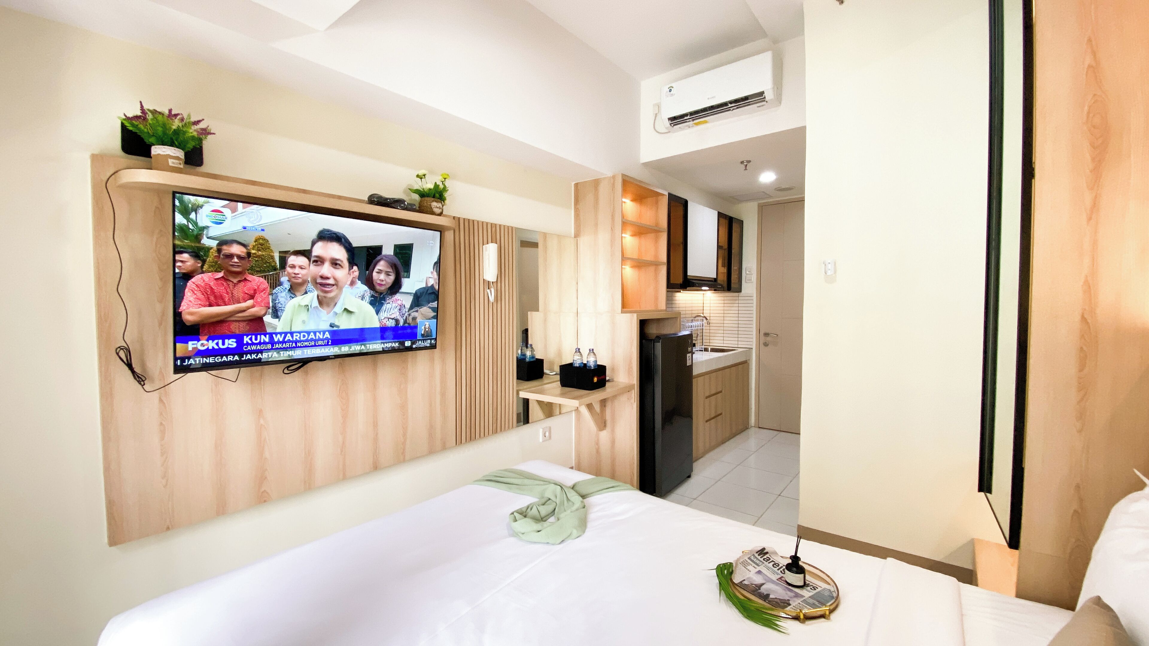 A hotel room with a TV mounted on the wall, a bed with a green towel, a small table with bottles, and a kitchen area with a refrigerator and sink.