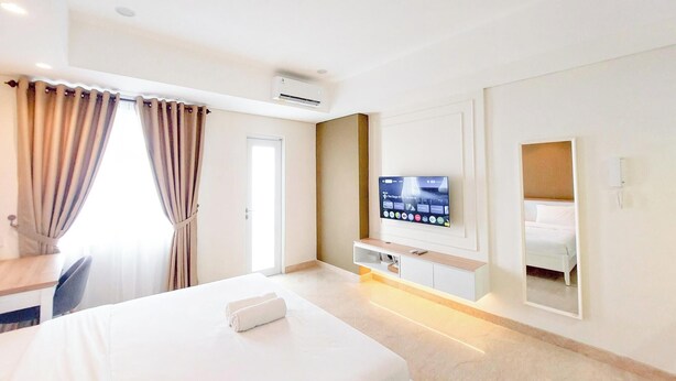 A hotel room with a bed, a TV mounted on the wall, a mirror, and a window with curtains. - Comfortable Studio At Podomoro City Deli Medan Apartment (Medan)