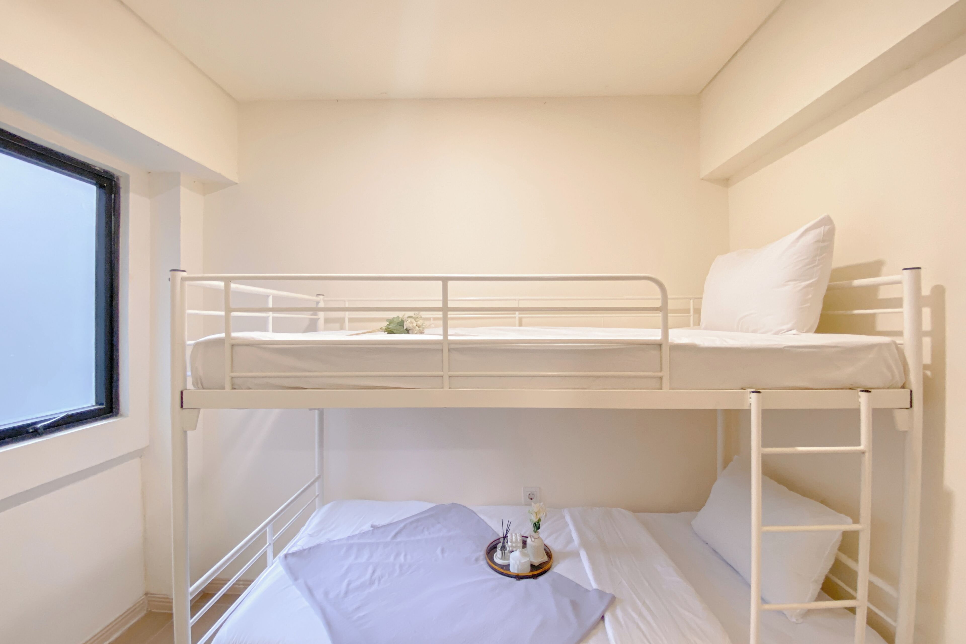 Habitación con literas, ropa de cama blanca, una mesita con un florero y velas, y una ventana a la izquierda.