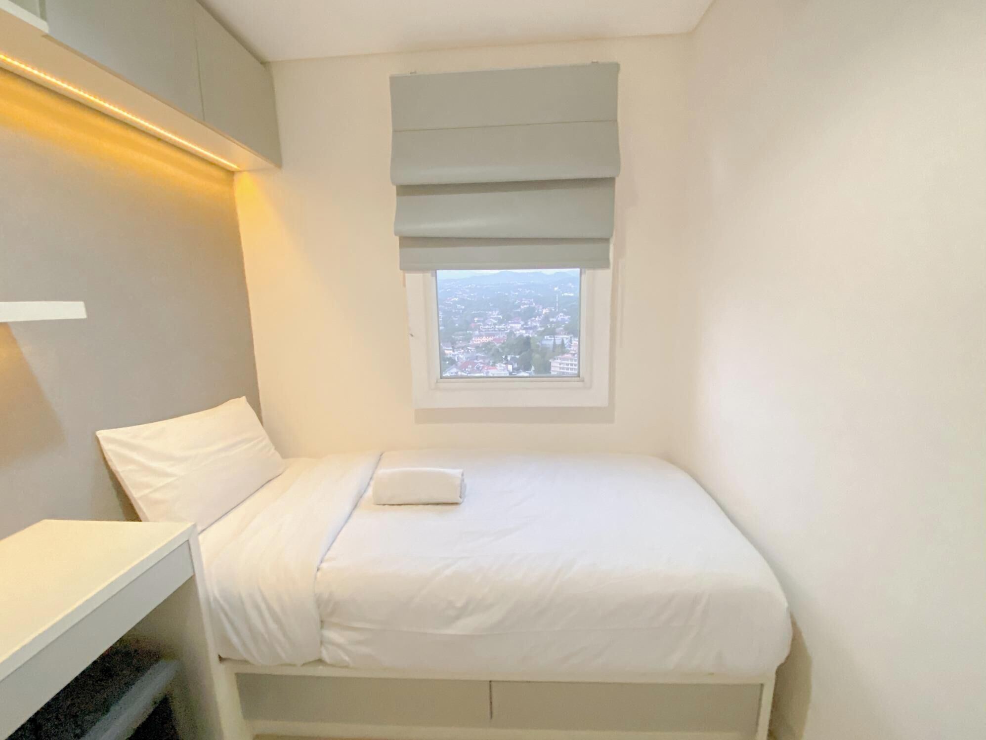 A small, neatly arranged bedroom with a single bed, a window with a view of a cityscape, and a built-in desk with storage.