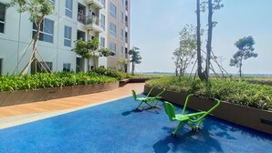 A rooftop pool area with blue flooring, green exercise chairs, and a view of a building with balconies and greenery. - Cozy And Great Deal Studio Apartment At Tokyo Riverside Pik 2 (Teluknaga)