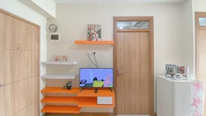 A compact living space with a TV on a wooden stand, a small shelf with decorative items, and a door with a mirror. - Good And Homey 2Br At Apartment Green Palm Residence (Jakarta)