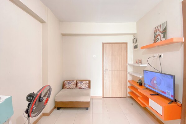 A small, well-lit room with a bed, a TV on an orange stand, a fan, and a door. - Good And Homey 2Br At Apartment Green Palm Residence (Jakarta)