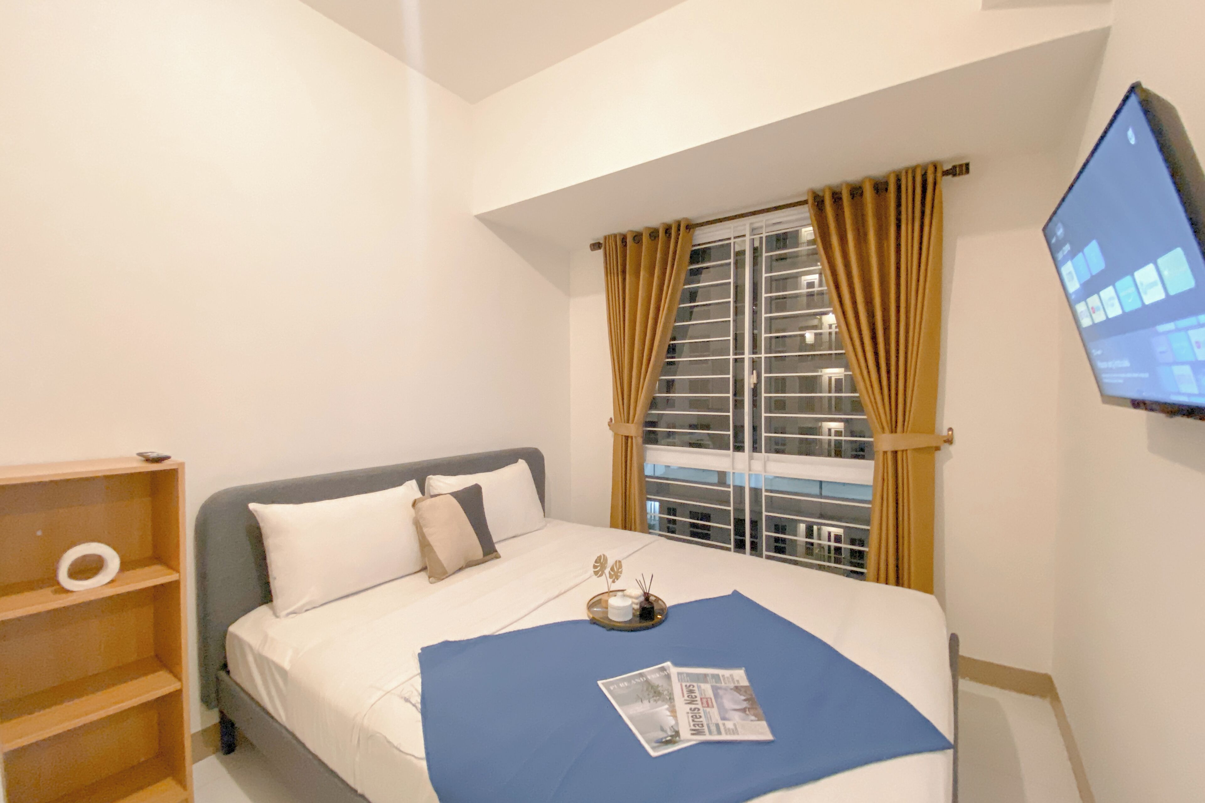 Un dormitorio con cama, ventana con cortinas, televisor en la pared y una repisa con un anillo.